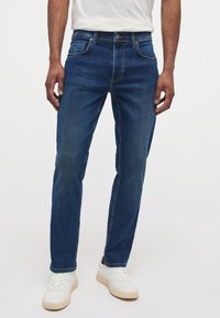 Jeanși din denim albastru cu o croială dreaptă, cinci buzunare și cusături contrastante. Asortați cu teniși albi, evidențiind un look casual.