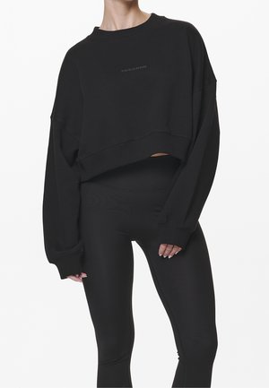 Frau trägt ein lockeres schwarzes Cropped-Sweatshirt und eng anliegende schwarze Leggings vor weißem Hintergrund.
