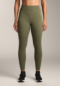 Nő olívazöld magasderekú leggingsben és fekete sportcipőben, semleges háttér előtt áll.