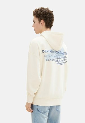 Sweat à capuche couleur crème avec un graphique au dos en bleu, portant le texte "DENIM & TOM TAILOR", des poignets côtelés et une poche kangourou. Porté avec un jean bleu clair.