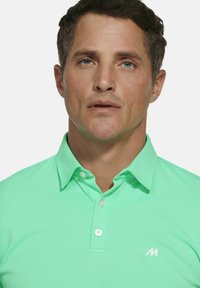 Lichtgroene polo met een gestructureerde stof, knoopsluiting en een klein wit logo op het linkerborstgebied.