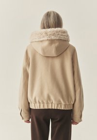 Beżowa kurtka z kapturem, z kołnierzem z futerka ekologicznego, elastycznym dołem i długimi rękawami. Tył wykonany jest z gładkiej tkaniny z subtelnymi przeszyciami.
