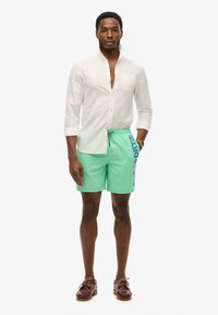 Costumi da bagno verde menta con cordoncino, con un logo blu sul lato, abbinati a una camicia di lino crema e mocassini marroni.