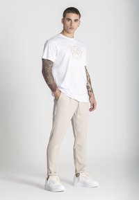 Biely tričko s jemným zlatým potlačou, v kombinácii s béžovými slim-fit nohavicami a bielymi teniskami s krémovými šnúrkami.