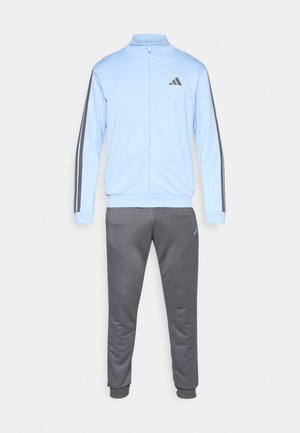 Svjetloplavi trenirka jakna s uspravnim okovratnikom i patentnim zatvaračem, u kombinaciji s tamnosivim hlačama; na rukavima su tri crne pruge i logotip.