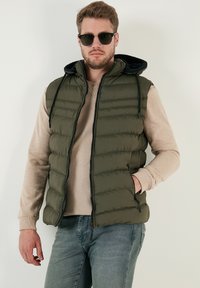 Olivgrön pufferväst med svart huva, dragkedjestängning och två sidofickor. Buren över en beige långärmad skjorta med mjuk textur.