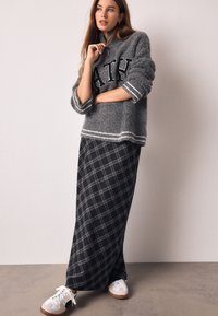 Pull en maille gris avec lettrage noir, associé à une longue jupe à carreaux noire et grise, complété par des baskets blanches.