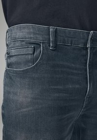 Gros plan de la taille et de la zone des poches d'un jean en denim bleu foncé porté avec une chemise noire rentrée.