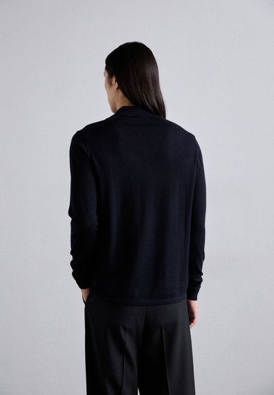 Schwarzer Pullover mit slim fit, langen Ärmeln, einer weichen Textur und einem dezenten Kragen. Kombiniert mit maßgeschneiderten schwarzen Hosen.