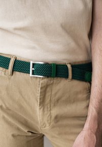 Cintura intrecciata verde con fibbia argentata e dettagli verdi, indossata sopra pantaloni beige. La cintura è realizzata in materiale flessibile con un motivo testurizzato.