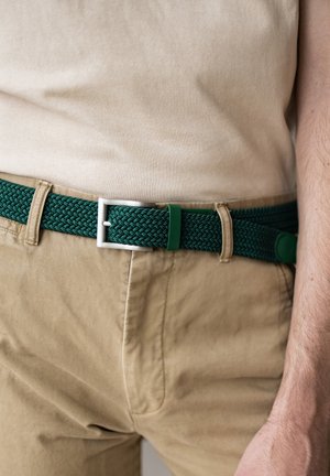 Ceinture tressée verte avec une boucle en argent et des accents verts, portée sur un pantalon beige. La ceinture est fabriquée en matériau flexible avec un motif texturé.