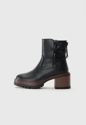 Bottine en cuir noir avec un talon épais en bois marron, semelle à crampons et une sangle décorative qui fait le tour à l'arrière.