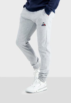 Pantalones de chándal grises de felpa con bolsillos laterales y un logo bordado, combinados con zapatillas altas blancas. El material es suave y ligeramente texturizado.