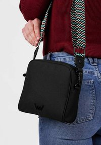 Černá crossbody taška vyrobená z odolné látky, s texturovaným popruhem s vzorem chevron a embosovaným logem na přední straně.