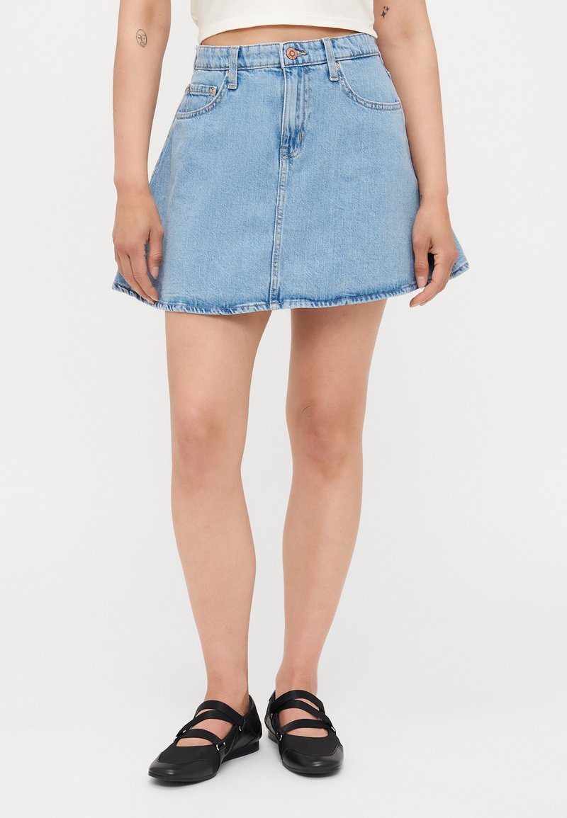 Femme portant une mini-jupe en jean bleu clair et des ballerines Mary Jane noires, debout, jambes légèrement écartées sur fond blanc.