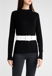 White leather belt with circular silver buckle worn over a black sweater, paired with black jeans. Sweater has a ribbed texture and crew neck.