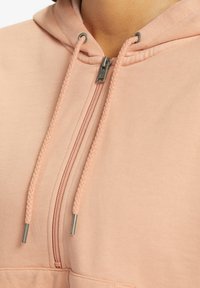 Sudadera color durazno con media cremallera en la parte delantera, cordones trenzados y cremallera metálica. Textura suave con un acabado sólido y liso.