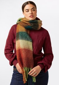 Écharpe en laine multicolore avec un dégradé de vert, d'orange et de marron, ornée de bords à franges, drapée sur un pull bordeaux.