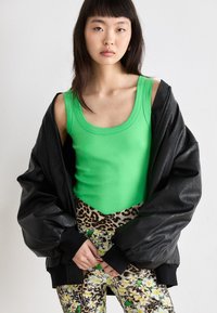 Zwarte leren oversized jas, felgroen tanktop en geprinte legging met luipaardmotief en bloemenaccenten. Matte textuur, felle kleuren.