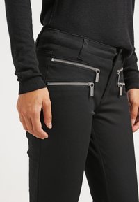 Pantalones negros con múltiples bolsillos con cremallera, hechos de un tejido suave. Presenta acentos de metal plateado y un diseño ajustado.