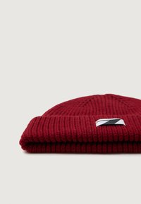 Gorro de punto rojo con un dobladillo, que presenta un parche de logo diagonal en blanco y negro en la parte frontal, con textura acanalada.
