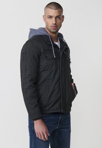 Melnā jaka ar pelēku kapuci, kurai ir vairāki priekšējie kabatas, rāvējslēdzis un ribotas aproces. Ikdieneša, izturīga auduma tekstūra.