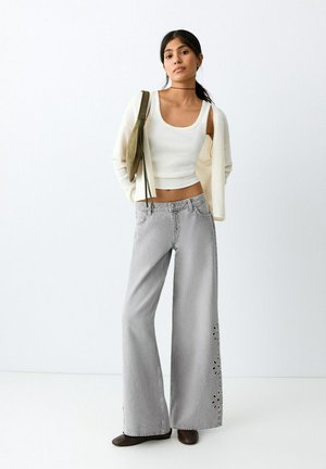 Joven mujer con camiseta blanca sin mangas, cárdigan crema, vaqueros grises de pierna ancha con detalles de flores recortadas, lleva un bolso de hombro oliva, de pie junto a una pared blanca.