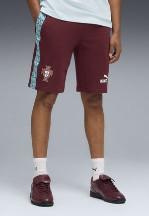 Man draagt bordeauxrode sportshorts met het voetbalwapen van Portugal en het Puma-logo, witte Puma-sokken en bordeauxrode sneakers, staand tegen een grijze achtergrond.
