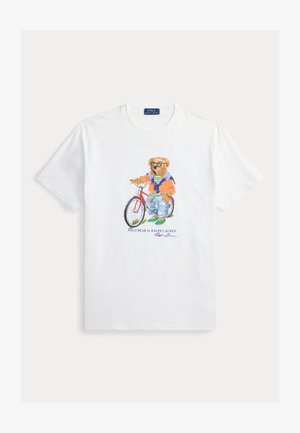 Balti marškinėliai trumpomis rankovėmis su meškiuku, dėvinčiu saulės akinius, džemperį, džinsus ir sportinius batelius, stovinčiu šalia raudono dviračio.