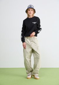Svart langermet skjorte med grafiske trykk på ermene, beige baggy bukser og en rutete bøttehatt, kombinert med lyse sneakers på et grønt gulv.