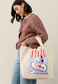 Plátená taška s grafikou homára, žiarivo červenými a modrými akcentmi a krémovým pozadím, ktorú drží osoba v textúrovanej hnedej kardigánke.