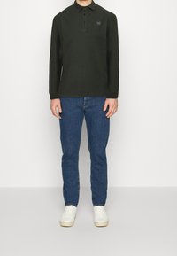 Camisa polo de manga larga en verde oscuro con un cierre de botones y un logotipo hexagonal. Combinada con jeans azules y zapatillas blancas. Diseño simple y casual.