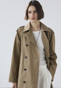 Brauner Trenchcoat mit doppelter Knopfleiste, großen Revers und schwarzen Knöpfen. Der Stoff wirkt glatt und leicht, mit Schulterklappen.
