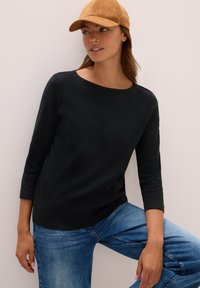 Frau mit einem hellbraunen Cap, schwarzem Langarmoberteil und blauen Jeans, die mit einem hochgezogenen Knie vor einer schlichten hellen Wand steht.