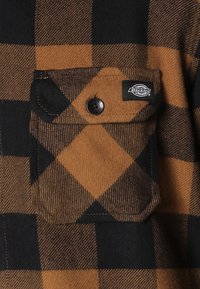 Tessuto di flanella a quadri marrone e nero con taschino sul petto dotato di bottone nero e etichetta con logo sulla tasca. Finitura texturizzata.