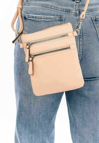 Světle růžová kožená crossbody taška se třemi zipovými kapsami a stříbrnými kovovými doplňky, zavěšená na odpovídajícím popruhu, vedle džínových kalhot.