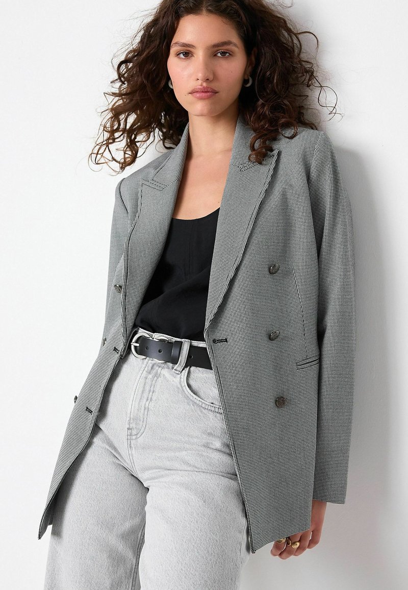 Double-breasted blazer in houndstooth pattern, gray and black. Features notched lapels, button accents, and a relaxed fit. Paired with gray jeans.