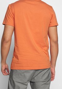 Camiseta de manga corta de color naranja, hecha de algodón, con un corte sencillo, que presenta un cuello redondo y una pequeña etiqueta en el dobladillo izquierdo.