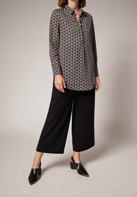 Blouse à manches longues boutonnée, avec un motif géométrique noir et crème, associée à un pantalon noir à jambes larges et des chaussures pointues foncées.