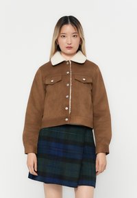 Jong vrouw in een bruine knopenjacket met een witte schapenvacht kraag en een blauw-groene geruite rok, staand tegen een witte achtergrond.