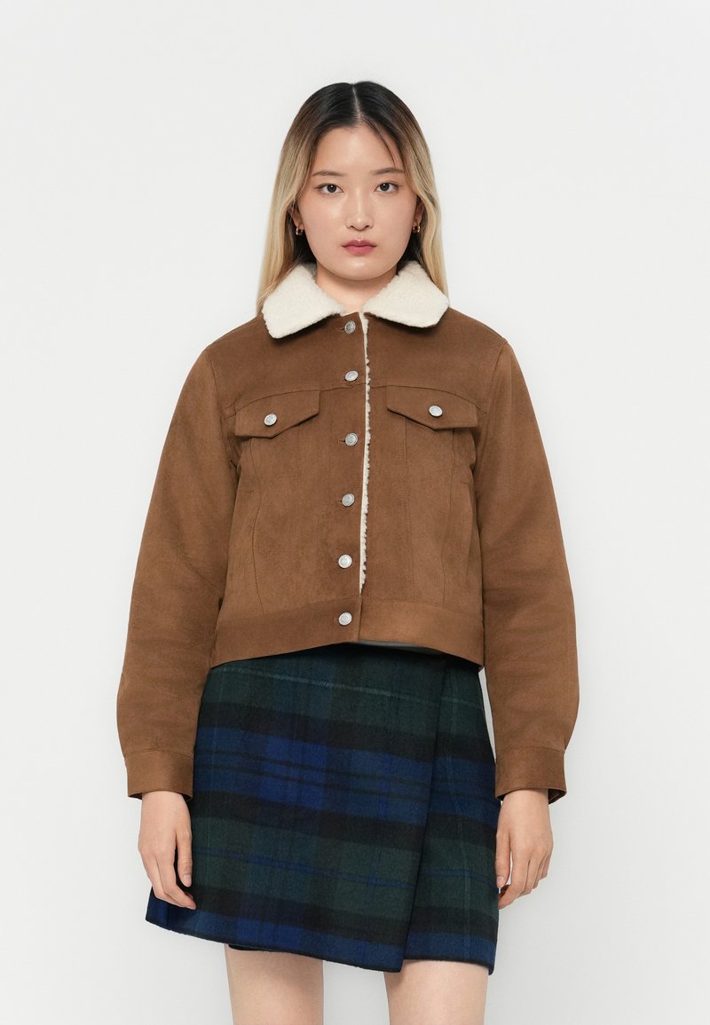 Jong vrouw in een bruine knopenjacket met een witte schapenvacht kraag en een blauw-groene geruite rok, staand tegen een witte achtergrond.