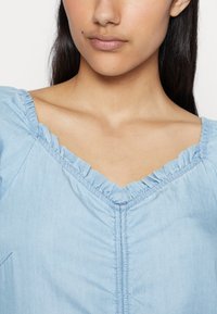 Top de algodão azul claro com decote em babados, design em forma de V e uma costura delicada a correr pelo centro. Textura suave.