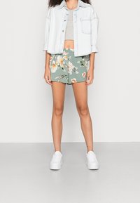 Veste en jean clair portée sur un crop top gris, associée à un short vert à motif floral. Des baskets blanches complètent la tenue, mettant en avant un look d'été décontracté.
