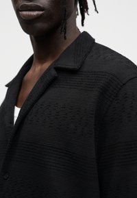 Μαύρο σακάκι με υφή και γιακά, με διακριτικό μοτίβο και κουμπιά. Φορεμένο πάνω από ένα λευκό εσώρουχο, με δομημένη εφαρμογή.