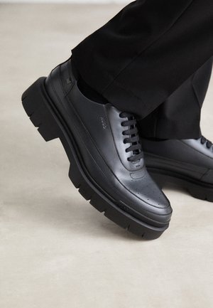 Czarne skórzane buty sznurowane z grubą masywną podeszwą noszone z czarnymi spodniami na jasnoszarej podłodze.