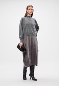 Γκρίζα πλεκτή μπλούζα με ψηλή λαιμόκοψη, layered πάνω από μια μεταξένια, μακριά, σκούρα γκρίζα φούστα. Η μοντέλα κρατά μια μαύρη τσάντα χεριού και φοράει μαύρες μπότες μέχρι το γόνατο.