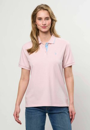 Femme aux longs cheveux blonds portant un polo rose clair à manches courtes et un jean bleu, debout devant un fond clair uni.