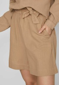 Lehká hnědá volně tkaná látková šortky s páskem na zavazování, bočními kapsami a uvolněným střihem. Vyznačují se hladkým povrchem a délkou ke kolenům.