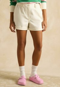 Pantaloni scurți albi din bumbac cu tivuri pliate, asortați cu saboți roz și șosete albe până la gleznă. Pantalonii scurți au în față un nasture și un fermoar.