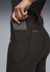 Χέρι που τοποθετεί ένα τηλέφωνο στην πλαϊνή τσέπη μαύρου κολάν Puma DryCell, με ουδέτερο φόντο.